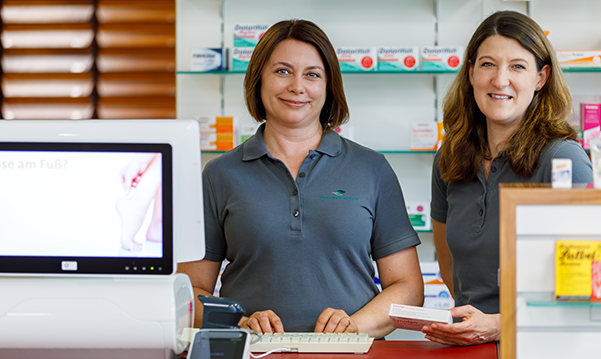 Zwei Apothekerin der Pluwig Apotheke stehen vor einem Medikamentenregal und arbeiten am Pc.
