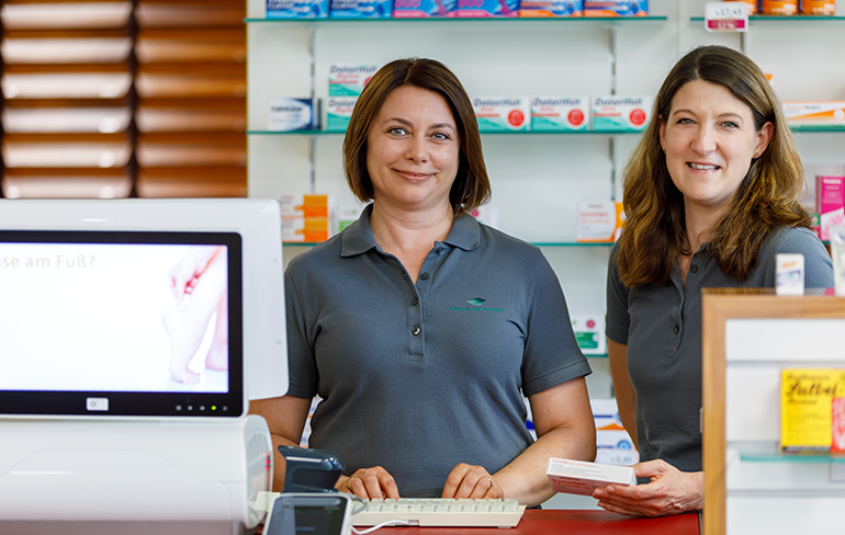 Zwei Mitarbeiterin der Pluwig Apotheke stehen vor dem Medikamentenregal am PC und halten ein Medikament in der Hand.