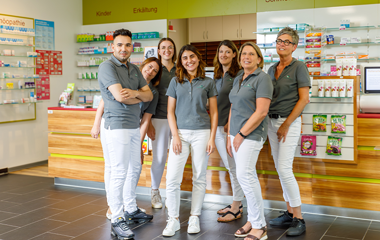 Das Team der Pluwig Apotheke in Pluwig steht in der Apotheke vor dem Tresen. Sie tragen grau Shirts mit dem Logo der Apotheke.