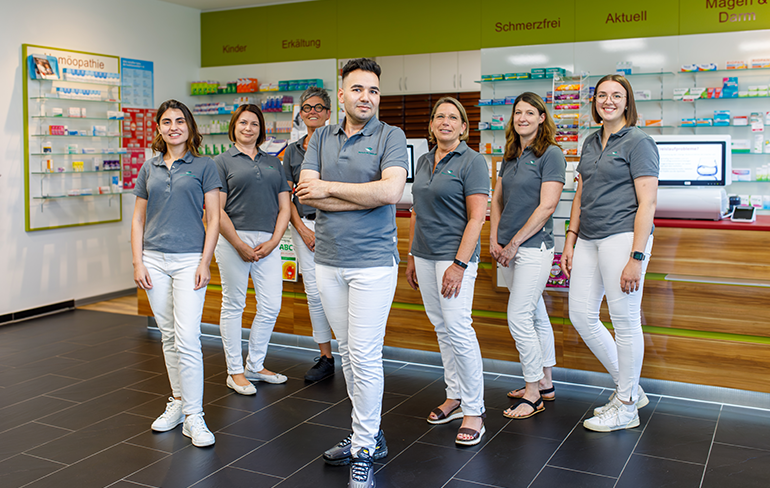 Das Team der Pluwig Apotheke in Pluwig steht in der Apotheke vor dem Tresen. Sie tragen grau Shirts mit dem Logo der Apotheke.