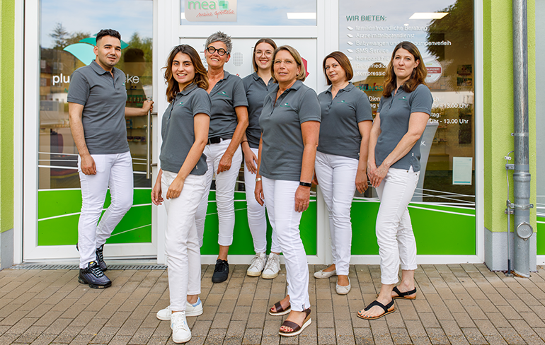 Das Team der Pluwig Apotheke in Pluwig steht vor der Apotheke vor dem Tresen. Sie tragen grau Shirts mit dem Logo der Apotheke.