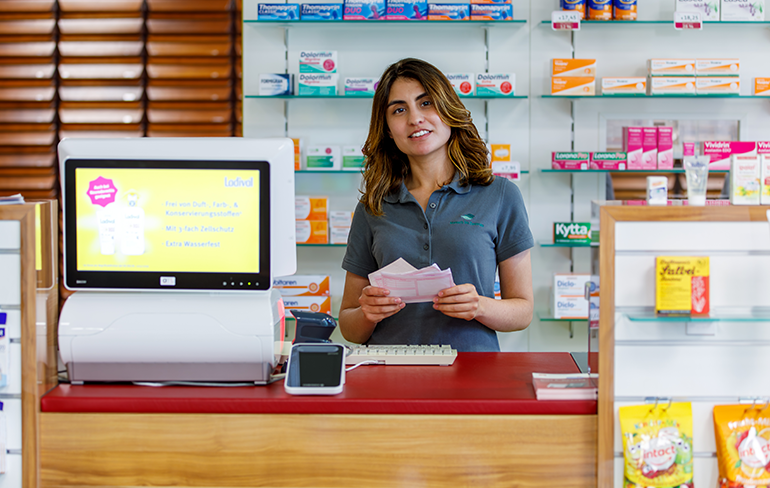 Eine Mitarbeiterin der Pluwig Apotheke steht vor dem Medikamentenregal und hält ein Rezept in der Hand.