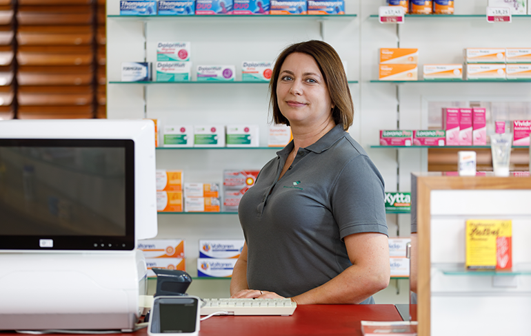 Eine Mitarbeiterin steht hinter dem Tresen am Pc der Pluwig Apotheke.
