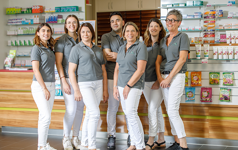 Das Team der Pluwig Apotheke in Pluwig steht in der Apotheke vor dem Tresen. Sie tragen grau Shirts mit dem Logo der Apotheke.