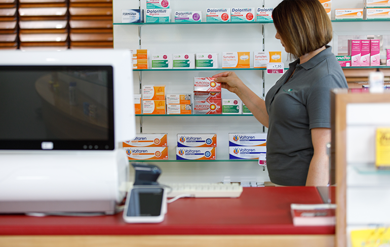 Eine Mitarbeiterin der Pluwig Apotheke stehen vor dem Medikamentenregal am PC und halten ein Medikament in der Hand.
