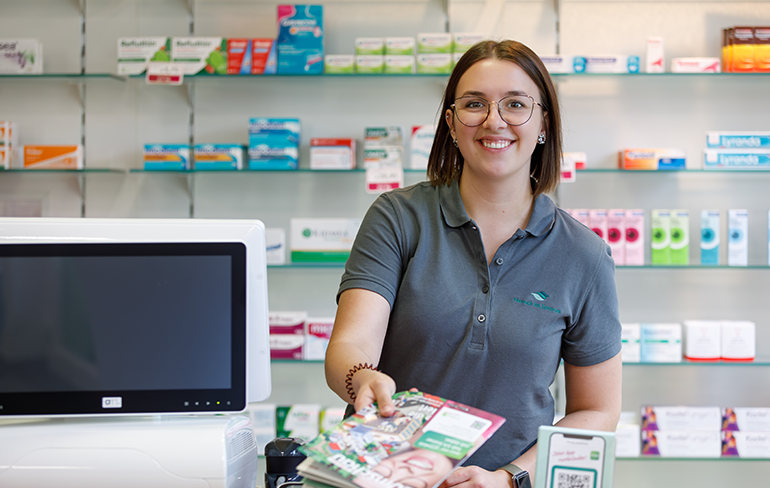 Eine Mitarbeiterin der Pluwig Apotheke steht vor dem Medikamentenregal und gibt ein Magazin über den Tresen.