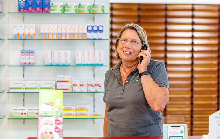 Eine Mitarbeiterin der Pluwig Apotheke steht vor dem Medikamentenregal der Pluwig Apotheke und telefoniert.