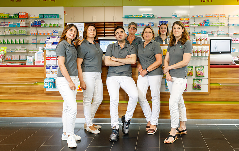 Das Team der Pluwig Apotheke in Pluwig steht in der Apotheke vor dem Tresen. Sie tragen grau Shirts mit dem Logo der Apotheke.