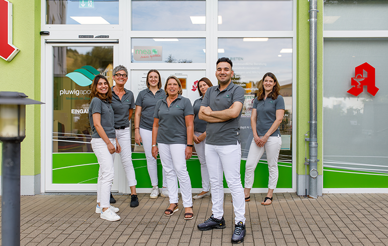 Das Team der Pluwig Apotheke in Pluwig steht vor der Tür der Apotheke. Sie tragen grau Shirts mit dem Logo der Apotheke.