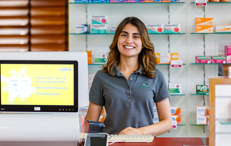 Eine Mitarbeiterin der Pluwig Apotheke steht vor dem Medikamentenregal am Pc.