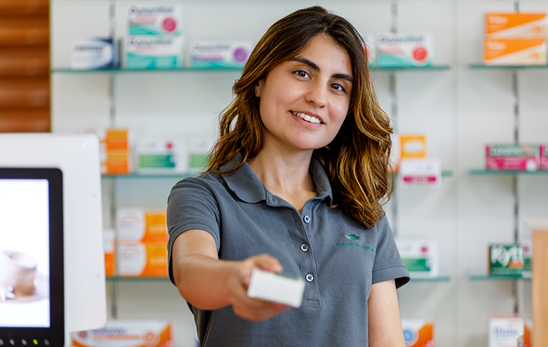 Eine Mitarbeiterin der Pluwig Apotheke steht vor dem Medikamentenregal und hält ein Medikament in der Hand.