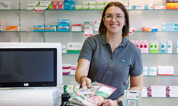 Apothekerin der Pluwig Apotheke in Pluwig steht hinter dem Tresen am Pc und reicht ein Magazin. Das unterstreicht die Fachkompetenz und technische Ausstattung der Apotheke.