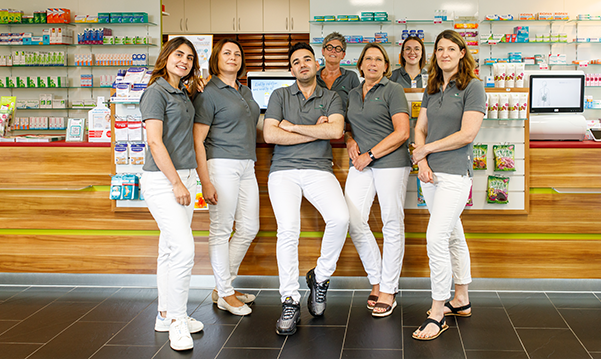 Das Team der Pluwig Apotheke in Pluwig steht in der Apotheke vor dem Tresen. Sie tragen grau Shirts mit dem Logo der Apotheke.