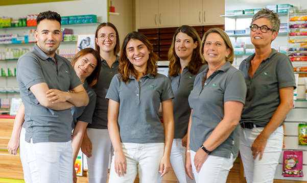 Das Team der Pluwig Apotheke in Pluwig steht in der Apotheke vor dem Tresen. Sie tragen grau Shirts mit dem Logo der Apotheke.